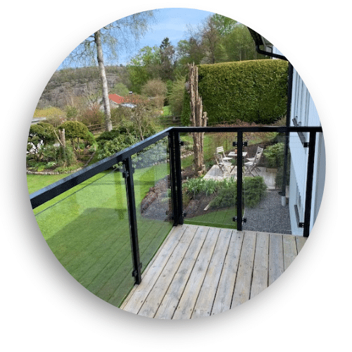 Elegant glassgjerde med svartlakkerte aluminiumsprofiler på terrasse med tretall, omgitt av frodig hage og grøntområde.