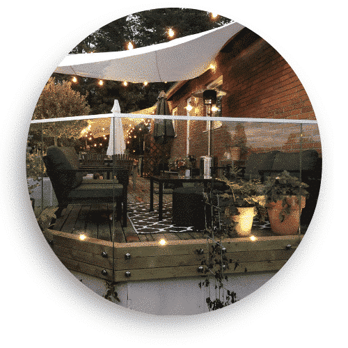 Altan med helglasräcke monterat på trästomme, omgiven av utemöbler, växter och stämningsfull belysning i trädgårdsmiljö.