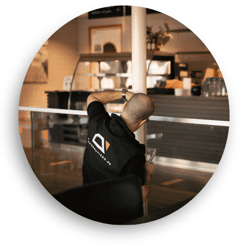 En montör i svart arbetsklädsel installerar ett glasräcke inomhus i en restaurangmiljö. Räcket har en minimalistisk design med rostfria beslag och skapar en stilren avskärmning mellan sittplatser och gångstråk.