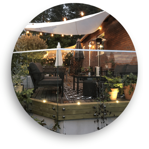 Glassrekkverk i kombinasjon av tre og glass på terrasse med kveldsbelysning, lyslenker og utemøbler.